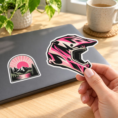 AI-generated illustration: An adult hand holds a pink-and-black helmet sticker beside a plain charcoal laptop with a pink mountain landscape sticker applied to its lid.